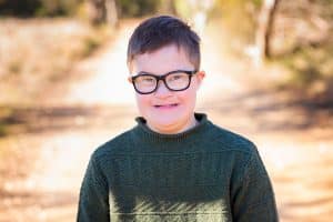 young boy with Down syndrome wearing black-rimmed glasses and a green sweater and smiling at the camera