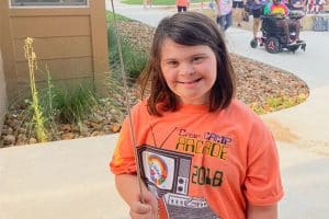 young girl with down syndrome at camp smiling at the camera and wearing an orange t-shirt
